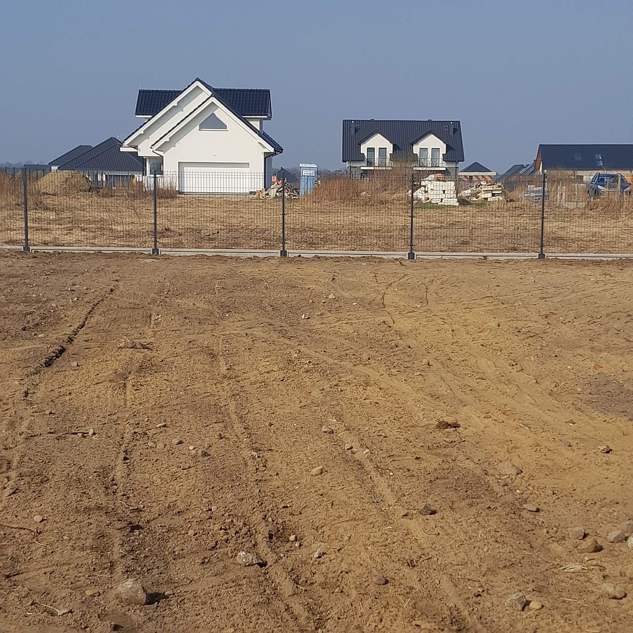 Nowoczesne ogrodzenie panelowe na tle nowo wybudowanych domów jednorodzinnych. Widok na brązową ziemię i czarne słupki ogrodzeniowe.