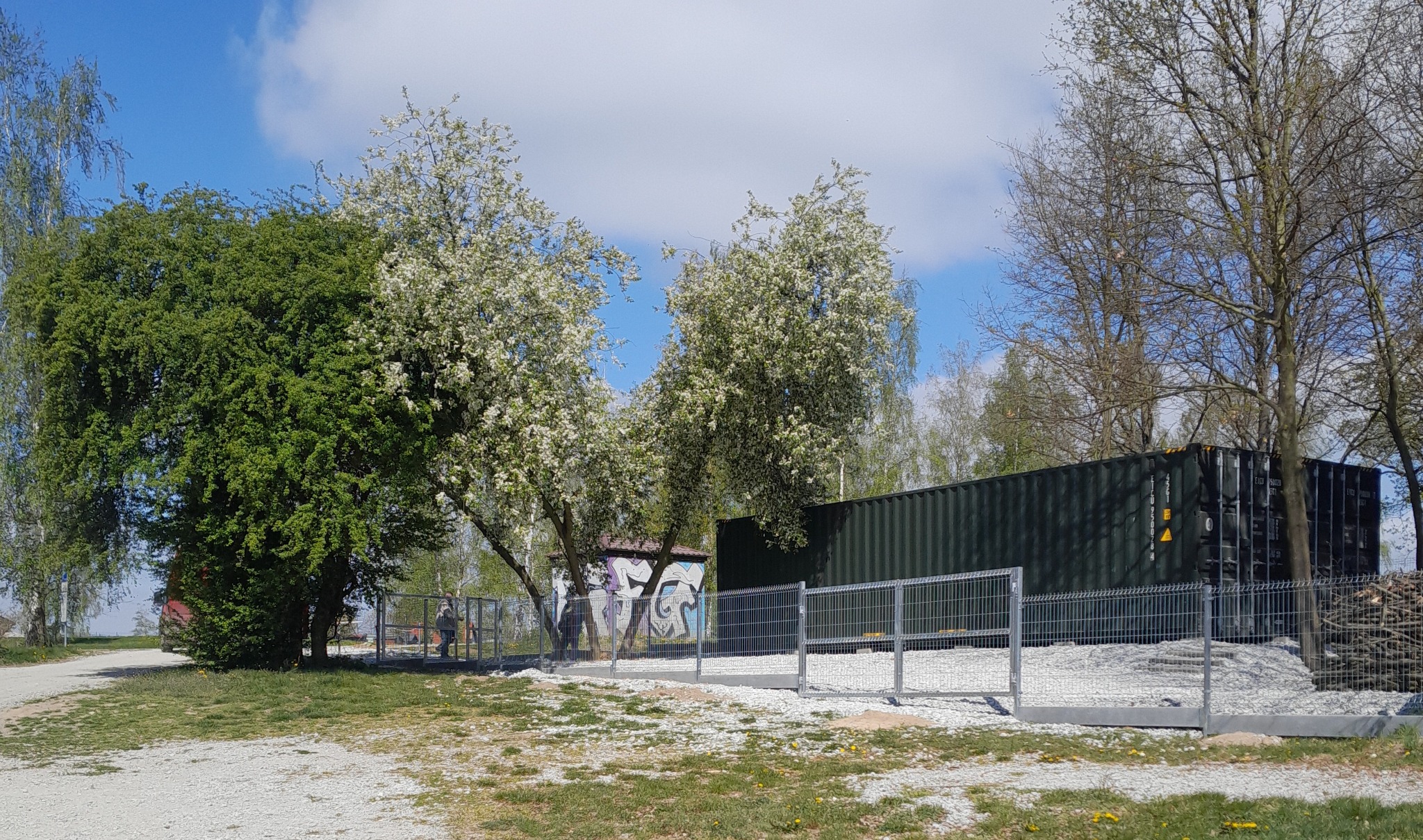 Nowoczesne ogrodzenie panelowe w kolorze szarym, otaczające teren z kontenerem. W tle drzewa i budynek z graffiti. Jasne niebo z chmurami. Widok z zewnątrz, wiosenna sceneria.
