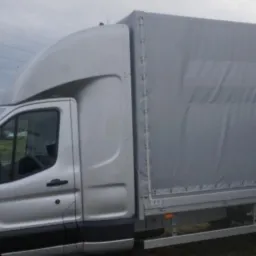 Srebrny samochód dostawczy z plandeką na tle parkingu i drzew bez liści.