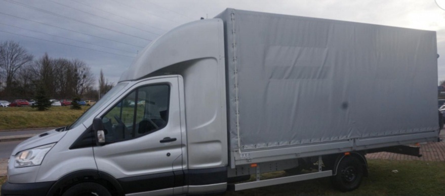 Srebrny samochód dostawczy z plandeką na tle parkingu i drzew bez liści.