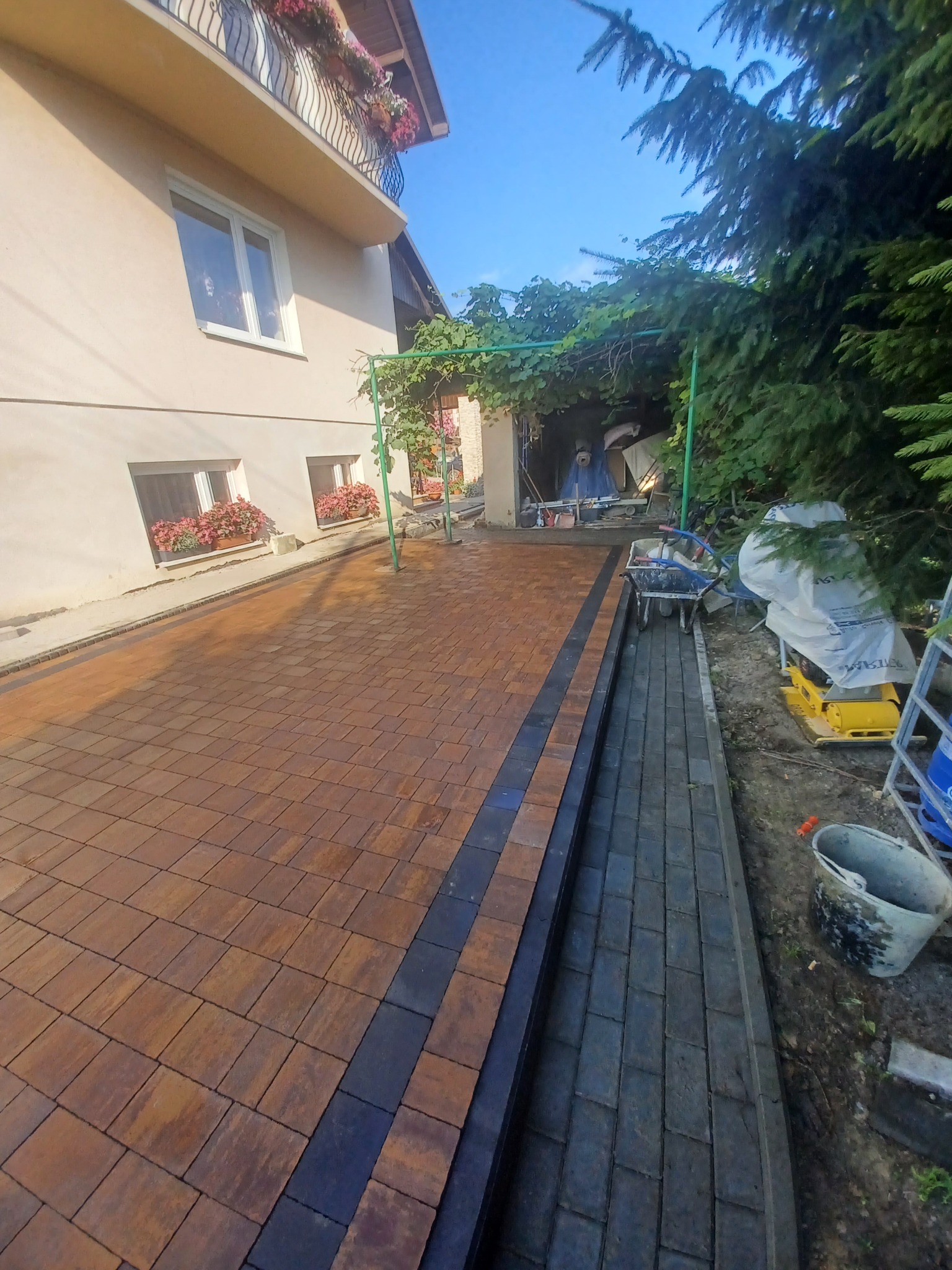 Nowo ułożona, brązowa kostka brukowa z czarnymi pasami, prowadząca do budynku z kwiatami w oknach i pod zadaszeniem, widoczny wózek budowlany i zagęszczarka.