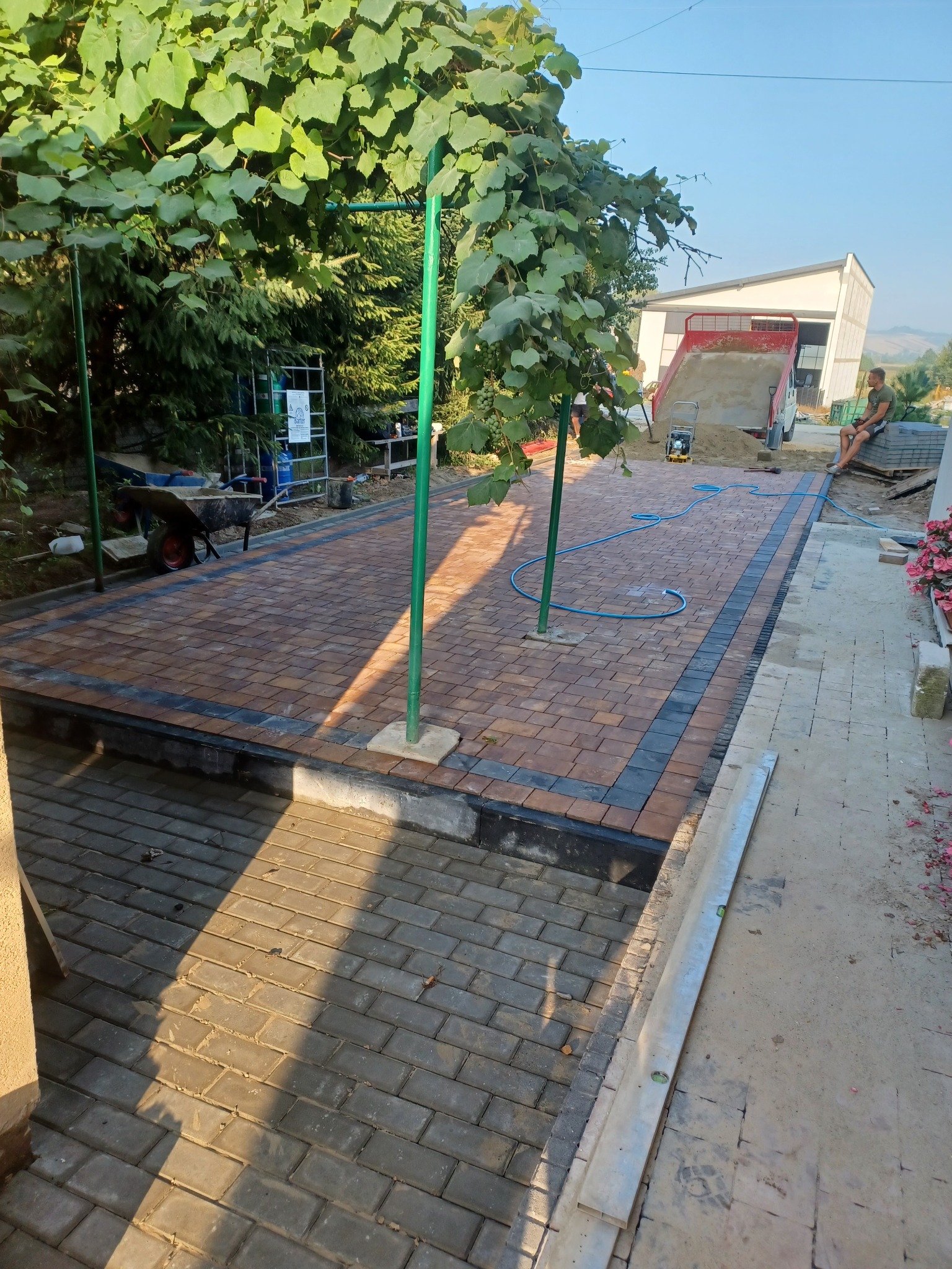 Ułożona kostka brukowa w ogrodzie z elementami zieleni, w tle wywrotka z piaskiem i osoba siedząca na palecie, widoczne narzędzia i węże.