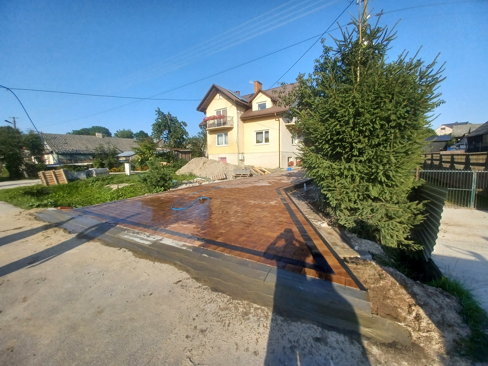 Świeżo ułożona, mokra kostka brukowa w odcieniach brązu, obrzeża z ciemnej kostki, w tle dom z balkonem i iglak, cień fotografa na pierwszym planie.