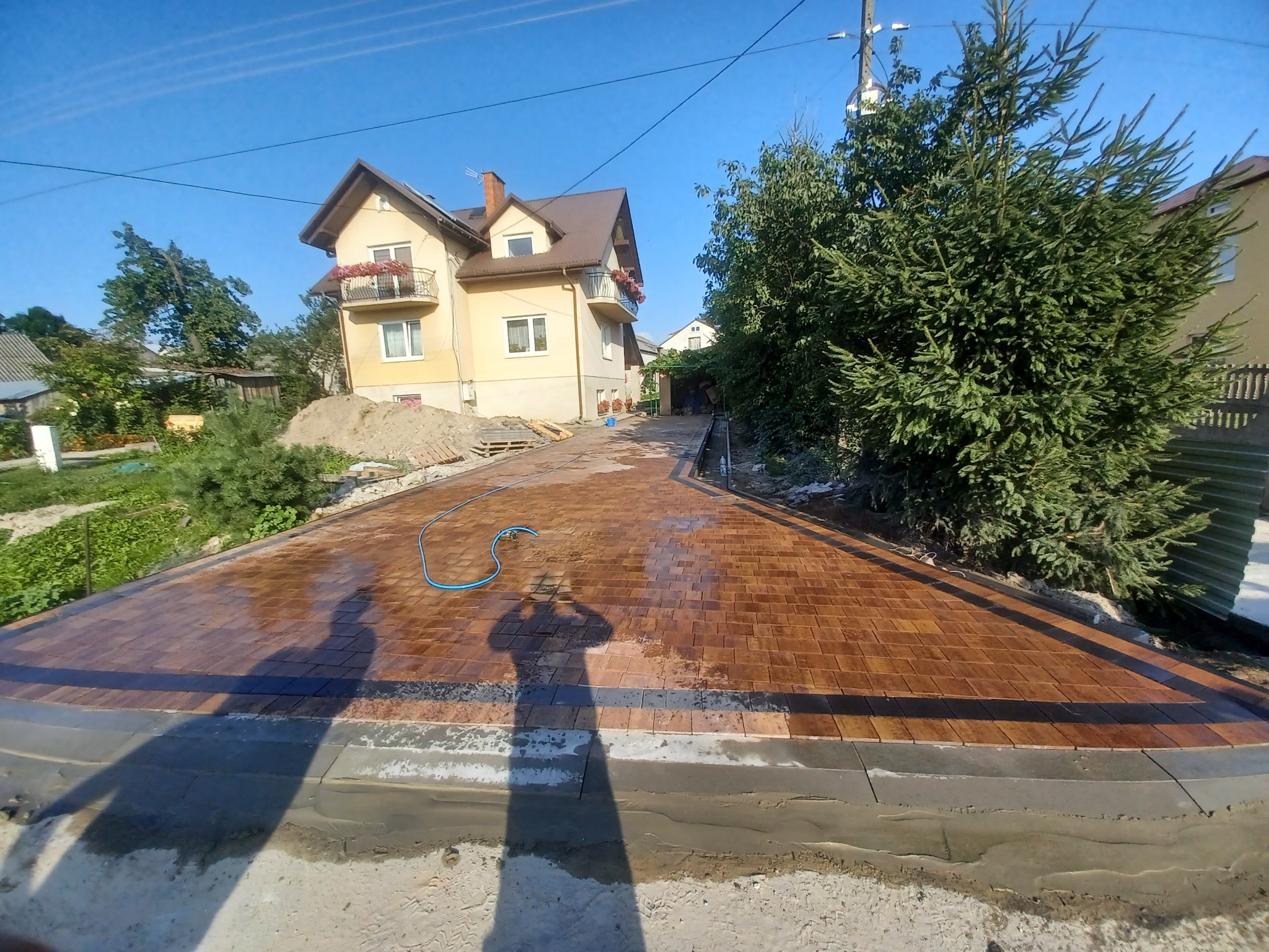 Nowo ułożona, mokra, brązowa kostka brukowa na podjeździe do domu jednorodzinnego z balkonami, widoczny cień osoby robiącej zdjęcie i niebieski wąż do podlewania.