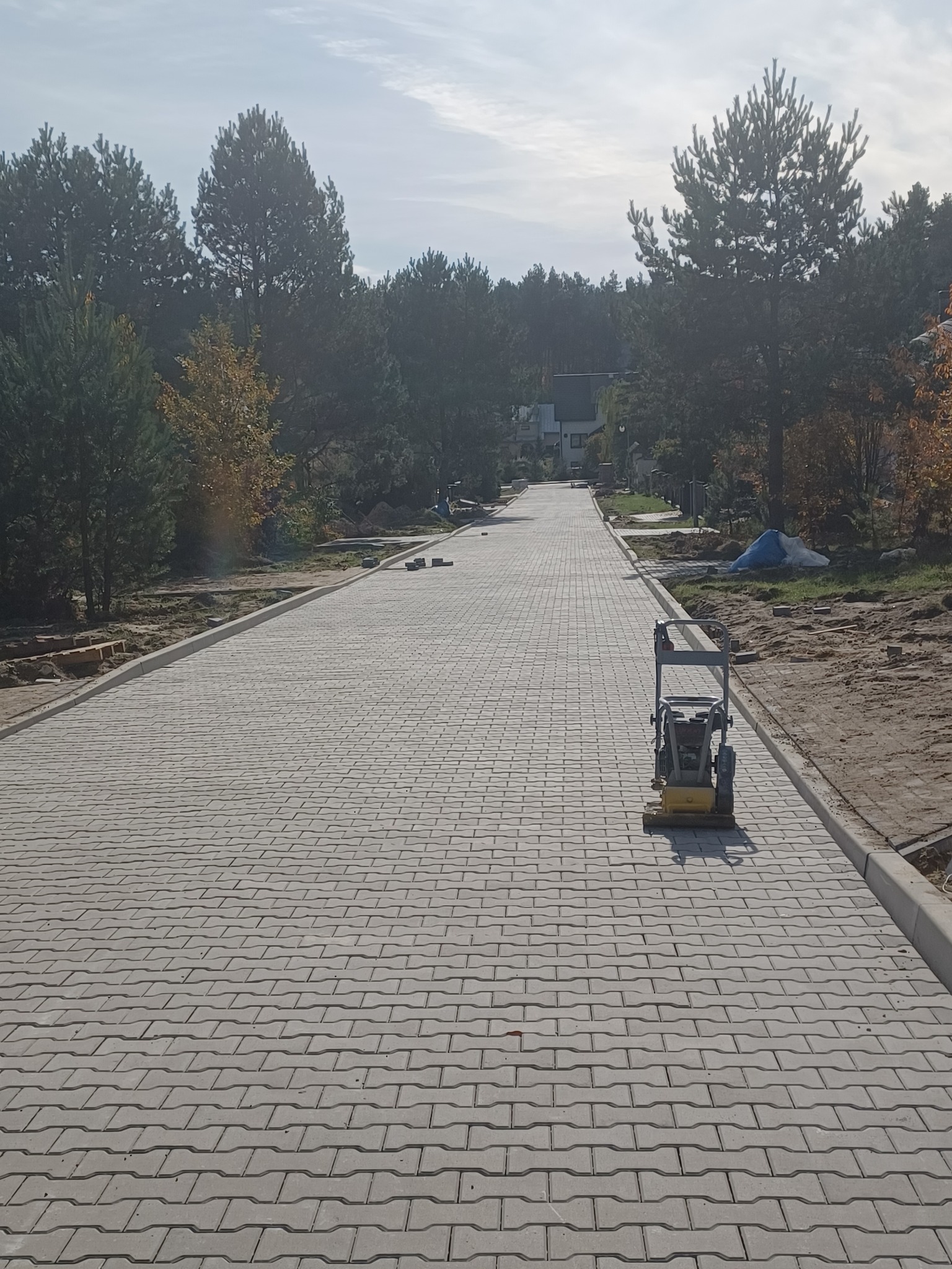 Długa, szara droga z kostki brukowej w trakcie budowy, z ubijarką wibracyjną na pierwszym planie i drzewami w tle.