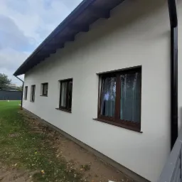 Boczny widok otynkowanego na biało domu jednorodzinnego z brązowymi oknami i czarną rynną, widoczne opadłe liście na trawie.