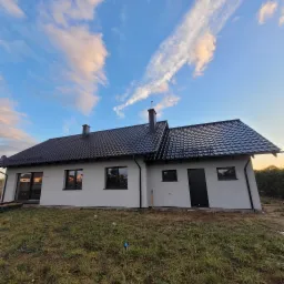 Nowoczesny dom jednorodzinny z ciemną dachówką i jasną elewacją, widok z dołu na tle błękitnego nieba z chmurami.