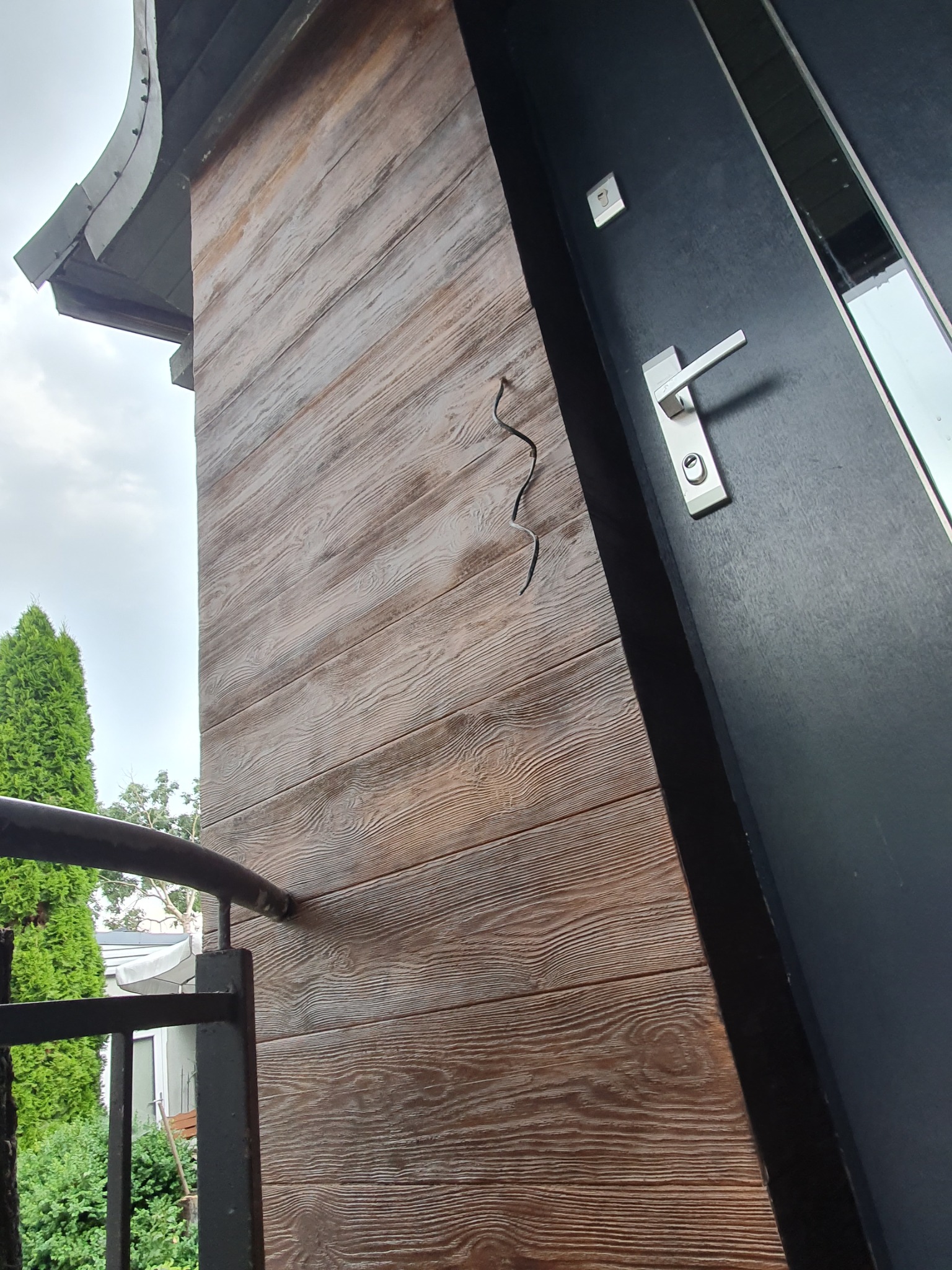 Fragment elewacji budynku z drewnopodobnym tynkiem w odcieniach brązu, obok czarne drzwi z metalową klamką, widoczny fragment balkonu i zieleni.