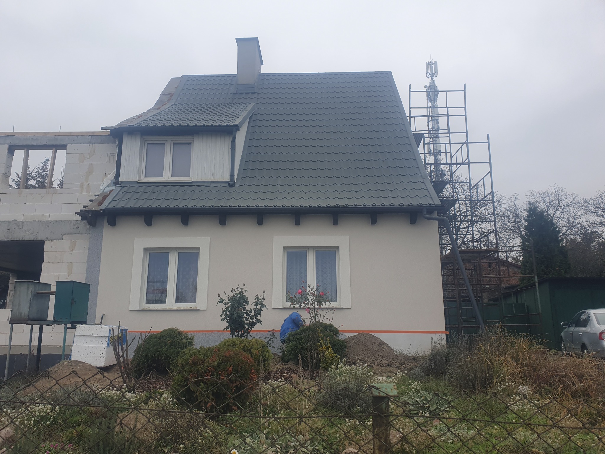 Dom jednorodzinny w trakcie prac wykończeniowych elewacji, z widocznym fragmentem nowej konstrukcji, rusztowaniem i elementami ocieplenia, widziany zza ogrodzenia.