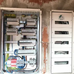 Dwie białe rozdzielnice elektryczne zamontowane na surowej ścianie z cegły. Lewa rozdzielnica otwarta, ukazująca liczne kable, bezpieczniki i listwy zaciskowe. Prawa rozdzielnica zamknięta, z logo...