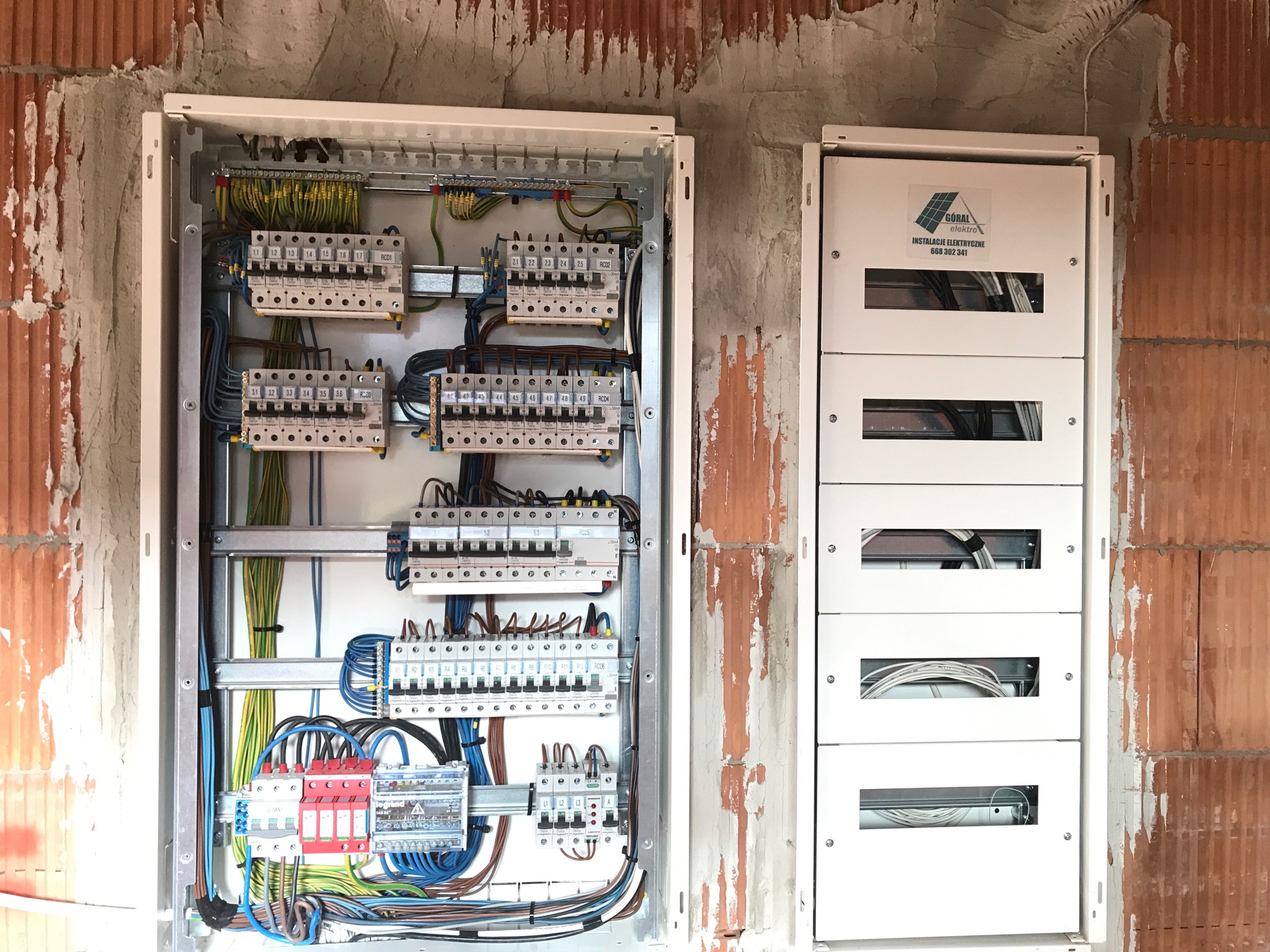 Dwie białe rozdzielnice elektryczne zamontowane na surowej ścianie z cegły. Lewa rozdzielnica otwarta, ukazująca liczne kable, bezpieczniki i listwy zaciskowe. Prawa rozdzielnica zamknięta, z logo...