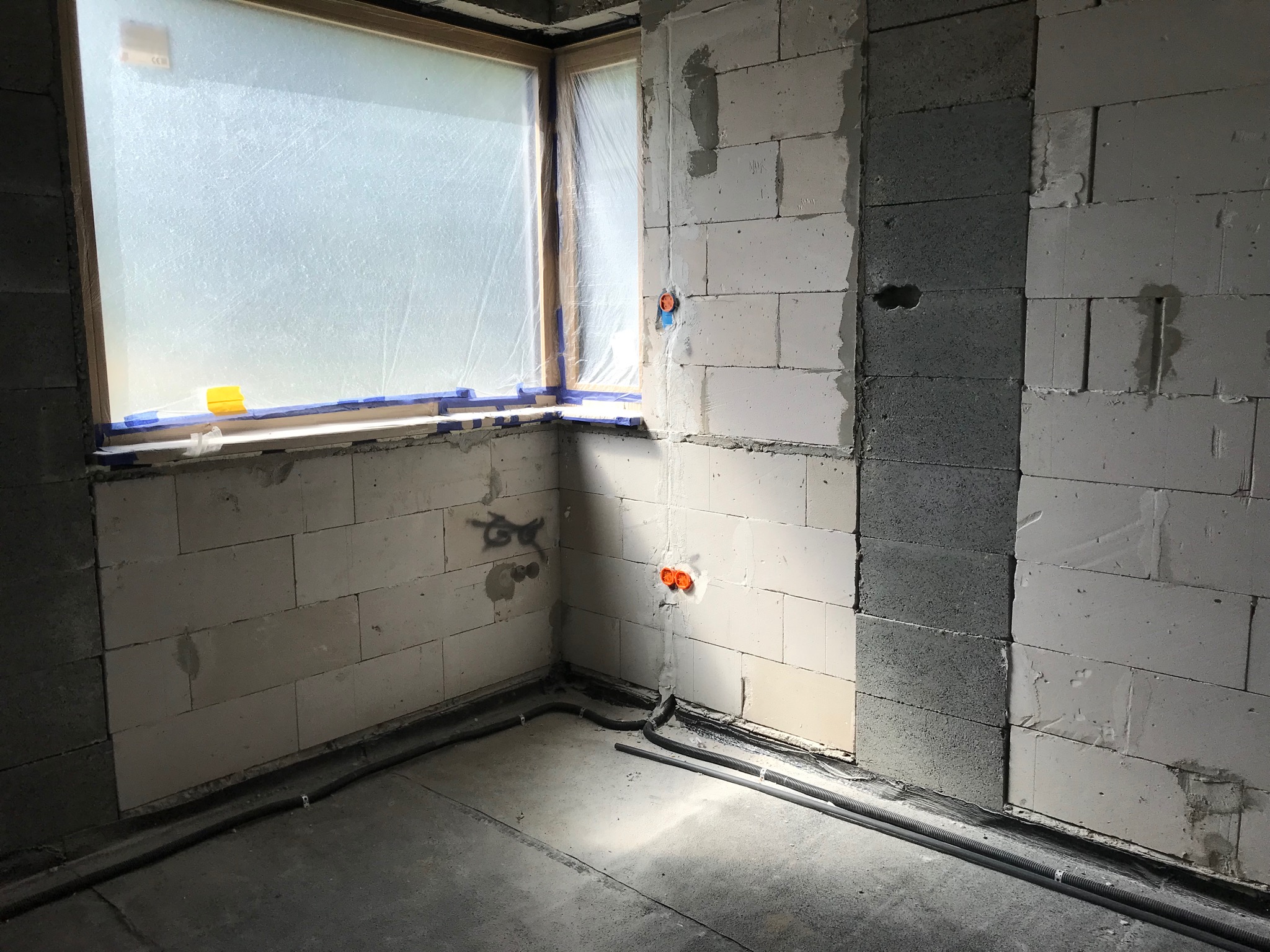 Surowy stan wnętrza z instalacją elektryczną w peszlach przy podłodze, widoczne puszki elektryczne w ścianie z betonu komórkowego, okno zabezpieczone folią malarską.