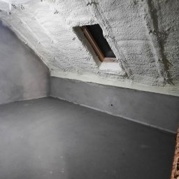 PMP-PUR Piotr Paczkowski - Poddasze po ociepleniu pianką: widoczne krokwie pokryte grubą warstwą pianki, okno dachowe z drewnianą ramą, ściany i podłoga w stanie surowym.