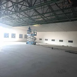 Pracownik na podnośniku koszowym wewnątrz hali z metalową konstrukcją dachu, przygotowujący się do prac izolacyjnych, widoczne wentylatory w ścianie