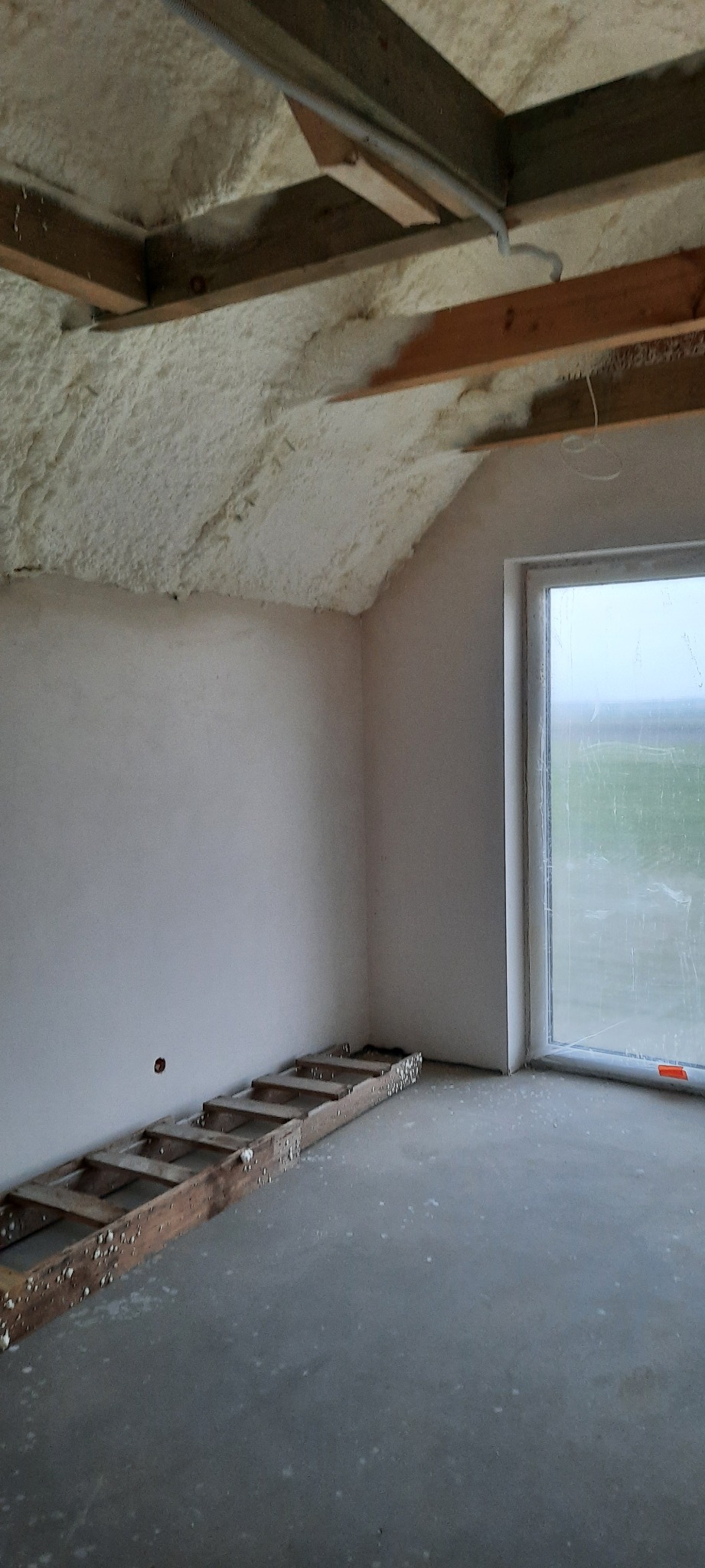 Poddasze po ociepleniu pianką: widoczne drewniane belki stropowe, biała pianka izolacyjna, fragment drabiny na podłodze, okno z widokiem na zewnątrz w pochmurny dzień.