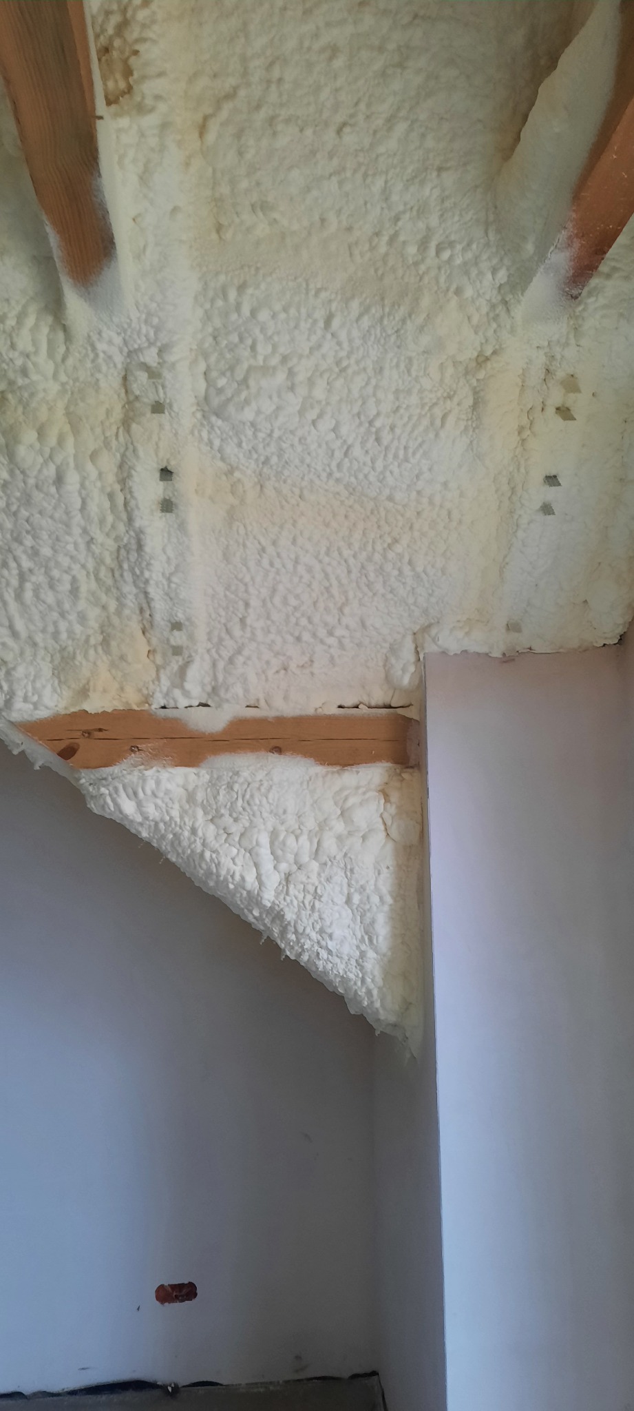 Poddasze z widoczną drewnianą konstrukcją i nałożoną grubą warstwą białej pianki izolacyjnej PUR, fragment ściany w jasnym kolorze, widoczne metalowe elementy mocujące.