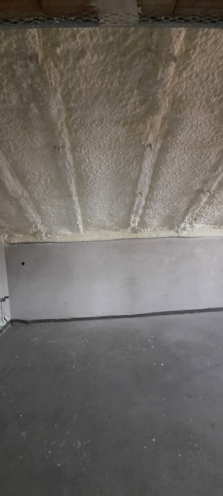 Poddasze po natrysku pianki poliuretanowej, widoczne mocowania na suficie, surowa ściana i betonowa podłoga.