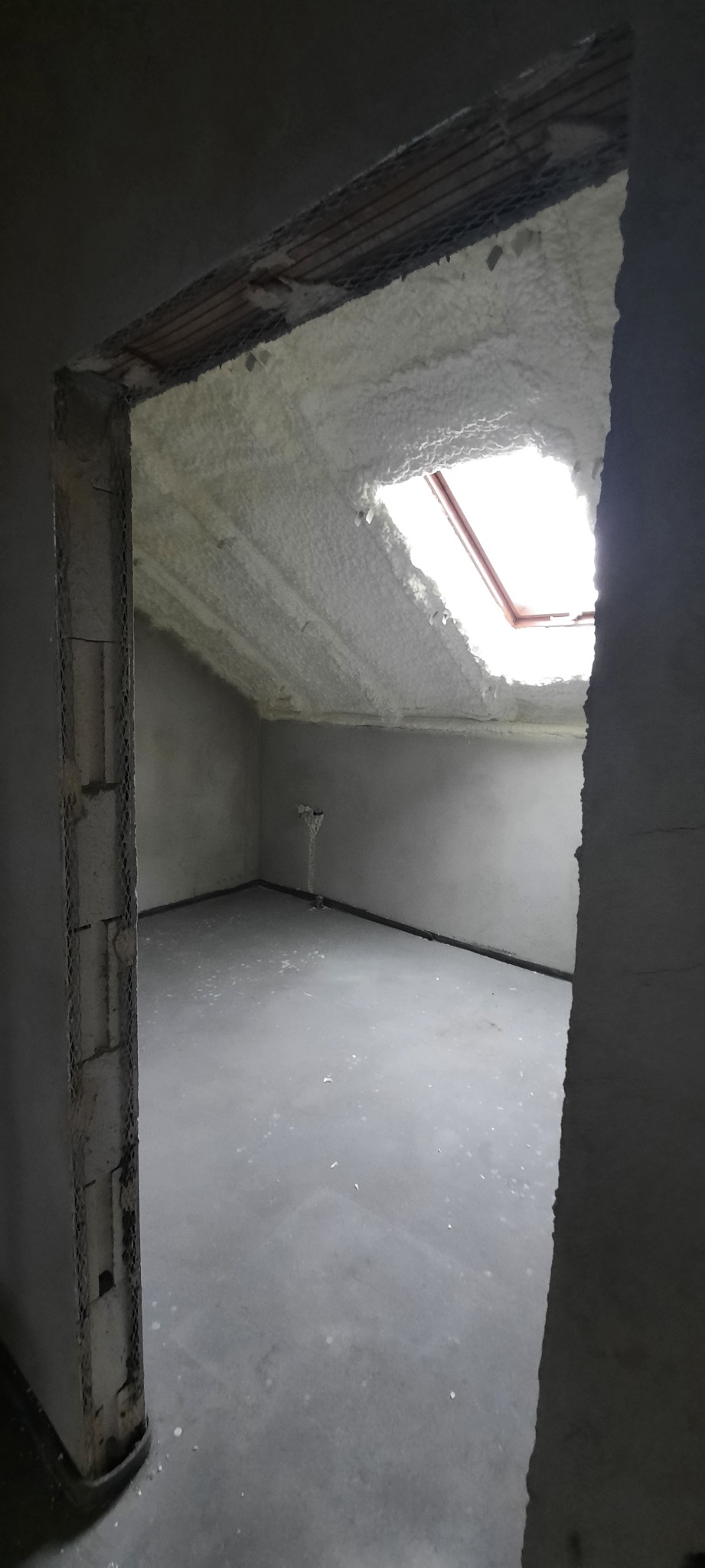 Poddasze z oknem dachowym po nałożeniu izolacji pianką, widok przez surowy otwór drzwiowy, szare ściany i podłoga w stanie deweloperskim.
