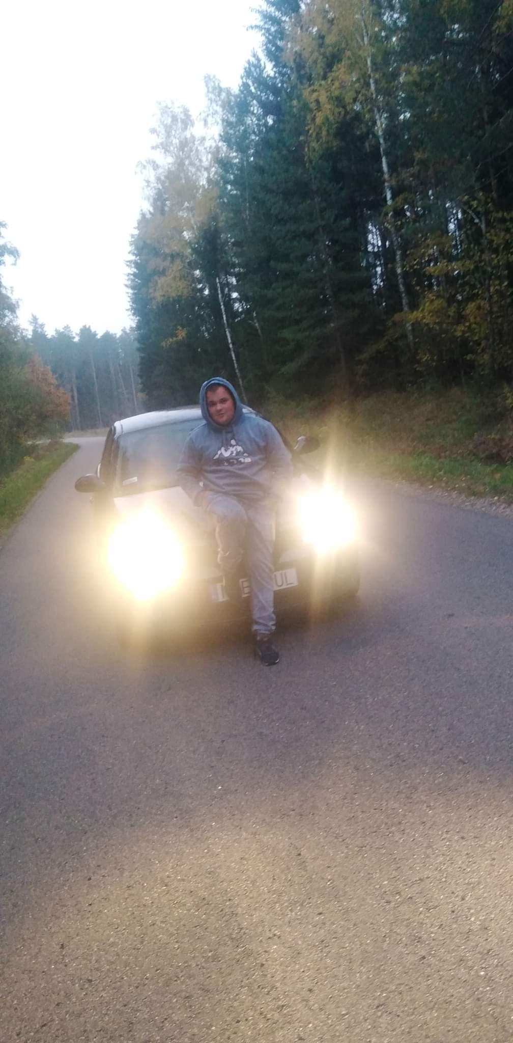 Młody mężczyzna w bluzie z kapturem opiera się o maskę samochodu na tle leśnej drogi, z włączonymi światłami.