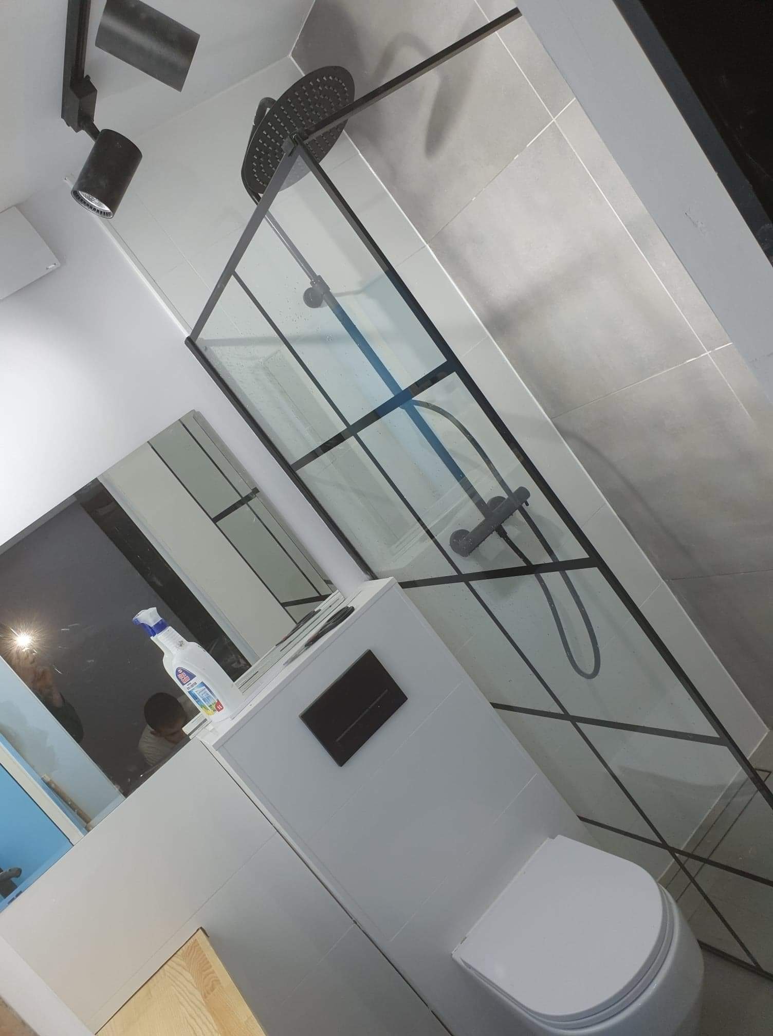 Nowoczesna kabina prysznicowa z czarnymi profilami i deszczownicą w łazience wykończonej szarymi płytkami, widoczny biały sedes i dozownik mydła na blacie umywalki.