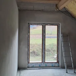 Wnętrze poddasza w trakcie prac wykończeniowych, ściany po nałożeniu tynku, widoczne okno z widokiem na zielone wzgórze, drabina oparta o ścianę.
