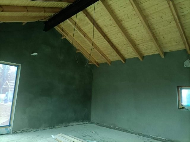 Wnętrze pomieszczenia w trakcie prac wykończeniowych; ściany pokryte świeżym, szarym tynkiem, drewniany strop z widocznymi belkami, surowe wykończenie podłogi, widoczne okno po lewej stronie.