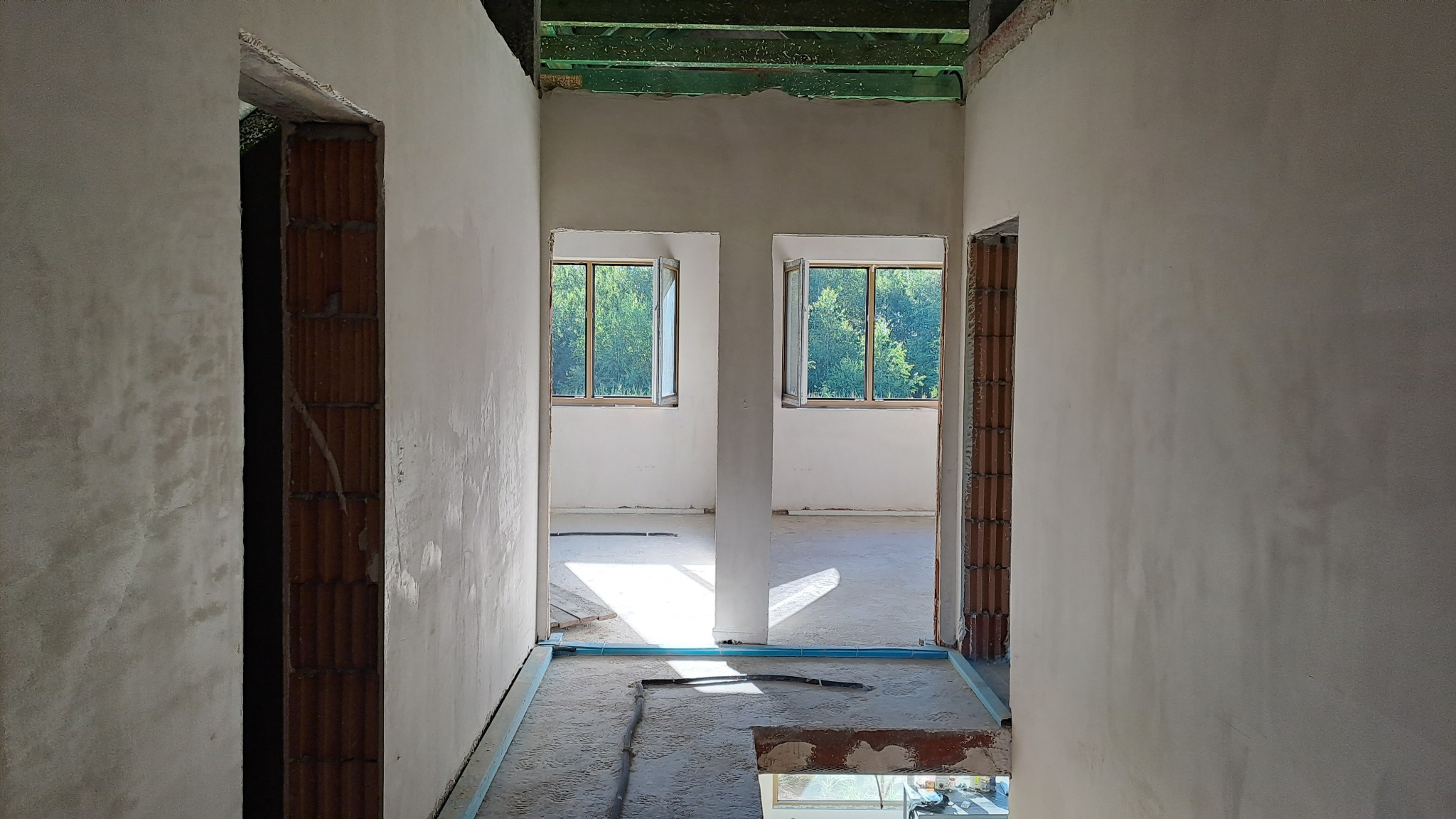 Surowe wnętrze z otynkowanymi ścianami w jasnym odcieniu szarości, widoczne fragmenty cegieł przy ościeżnicach, nieukończona podłoga i dwa okna z widokiem na zieleń.