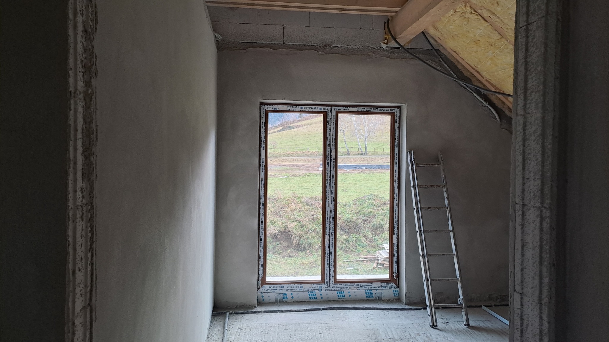 Wnętrze poddasza w trakcie prac wykończeniowych, ściany po nałożeniu tynku, widoczne okno z widokiem na zielone wzgórze, drabina oparta o ścianę.