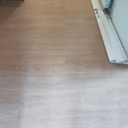 Jasna podłoga drewnopodobna w trakcie remontu, listwy przypodłogowe ułożone wzdłuż ściany, widoczny grzejnik i narzędzia malarskie w tle.