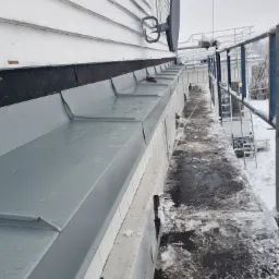 Nowa, szara rynna zamontowana na białej ścianie budynku z widocznym fragmentem dachu i anteną satelitarną, sfotografowana z rusztowania w zimowy dzień.