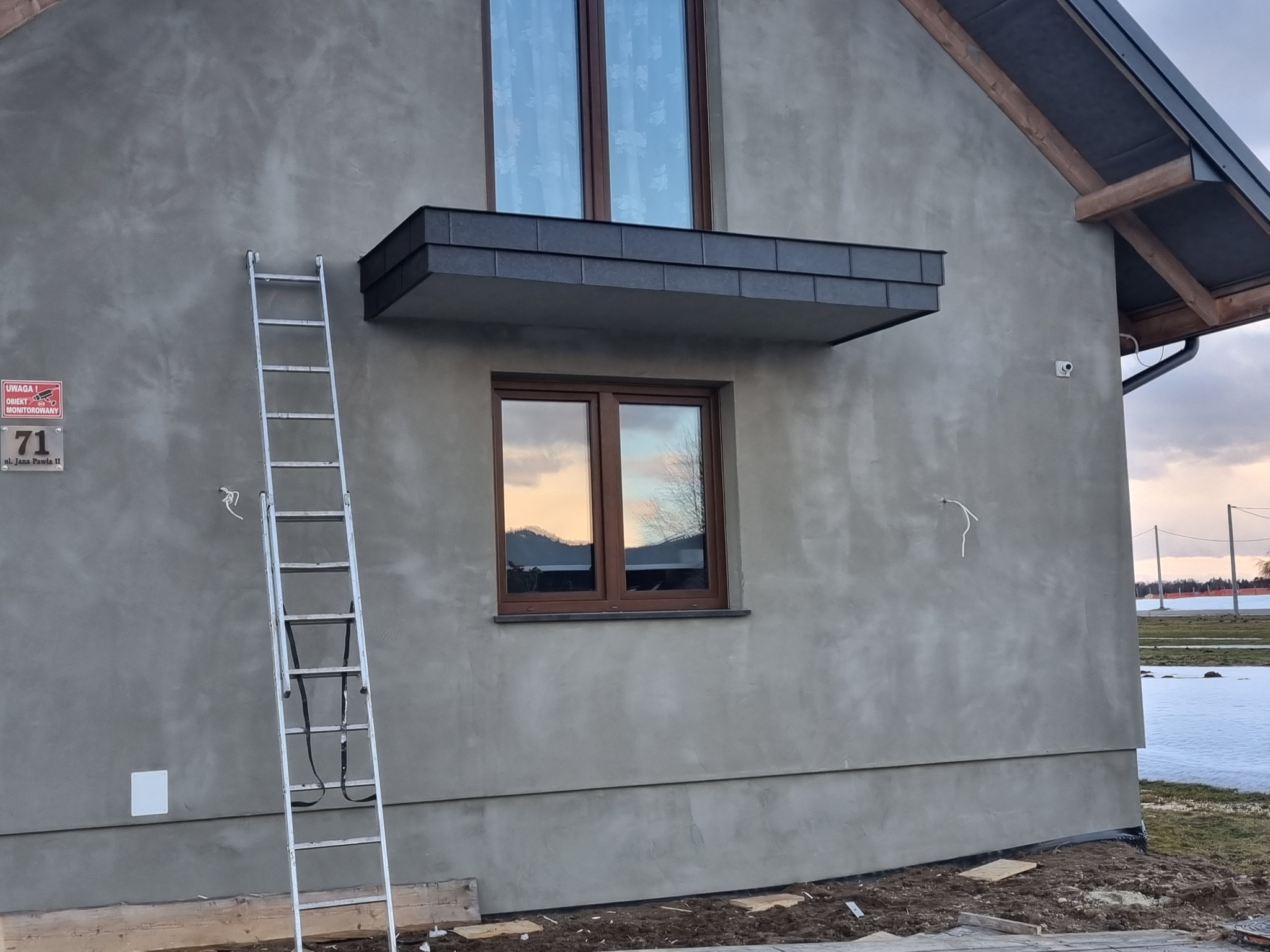 Świeżo otynkowana ściana domu jednorodzinnego z zamontowanym balkonem francuskim z szarych płytek, widoczna aluminiowa drabina oparta o ścianę, częściowo ośnieżony krajobraz w tle.