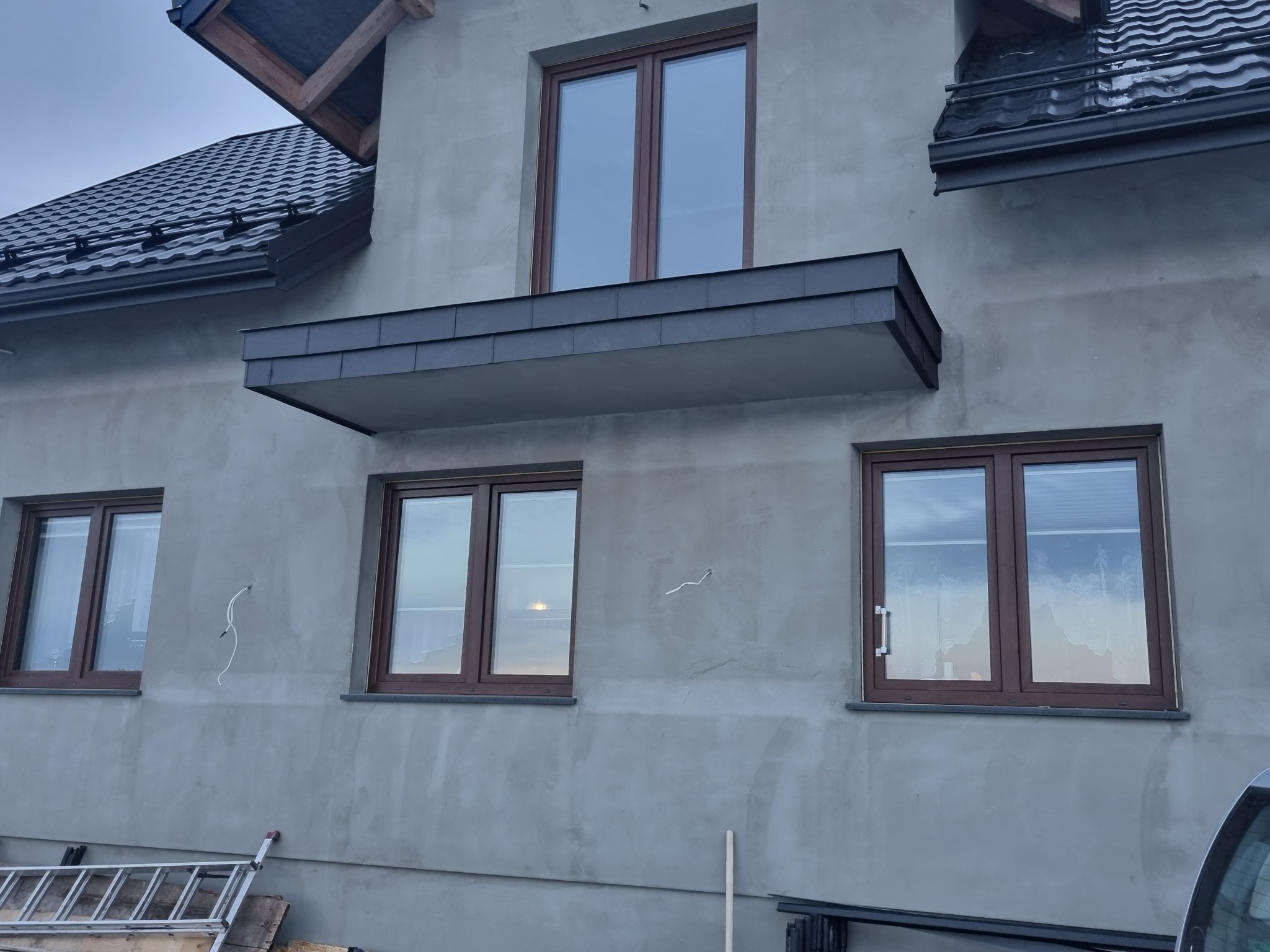 Surowy stan elewacji domu jednorodzinnego z zamontowanymi oknami w brązowych ramach, widoczne wystające przewody i fragment rusztowania, czarny dach z blachy trapezowej.