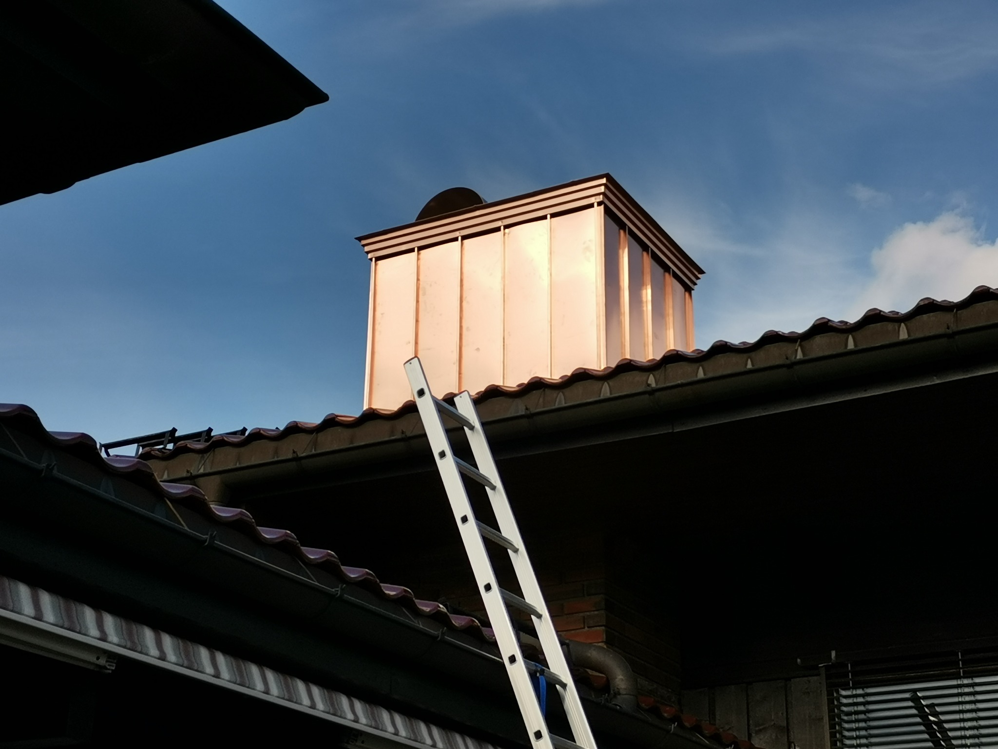 Miedziany komin na dachu z drabina opartą o rynnę, widok z dołu na tle błękitnego nieba.