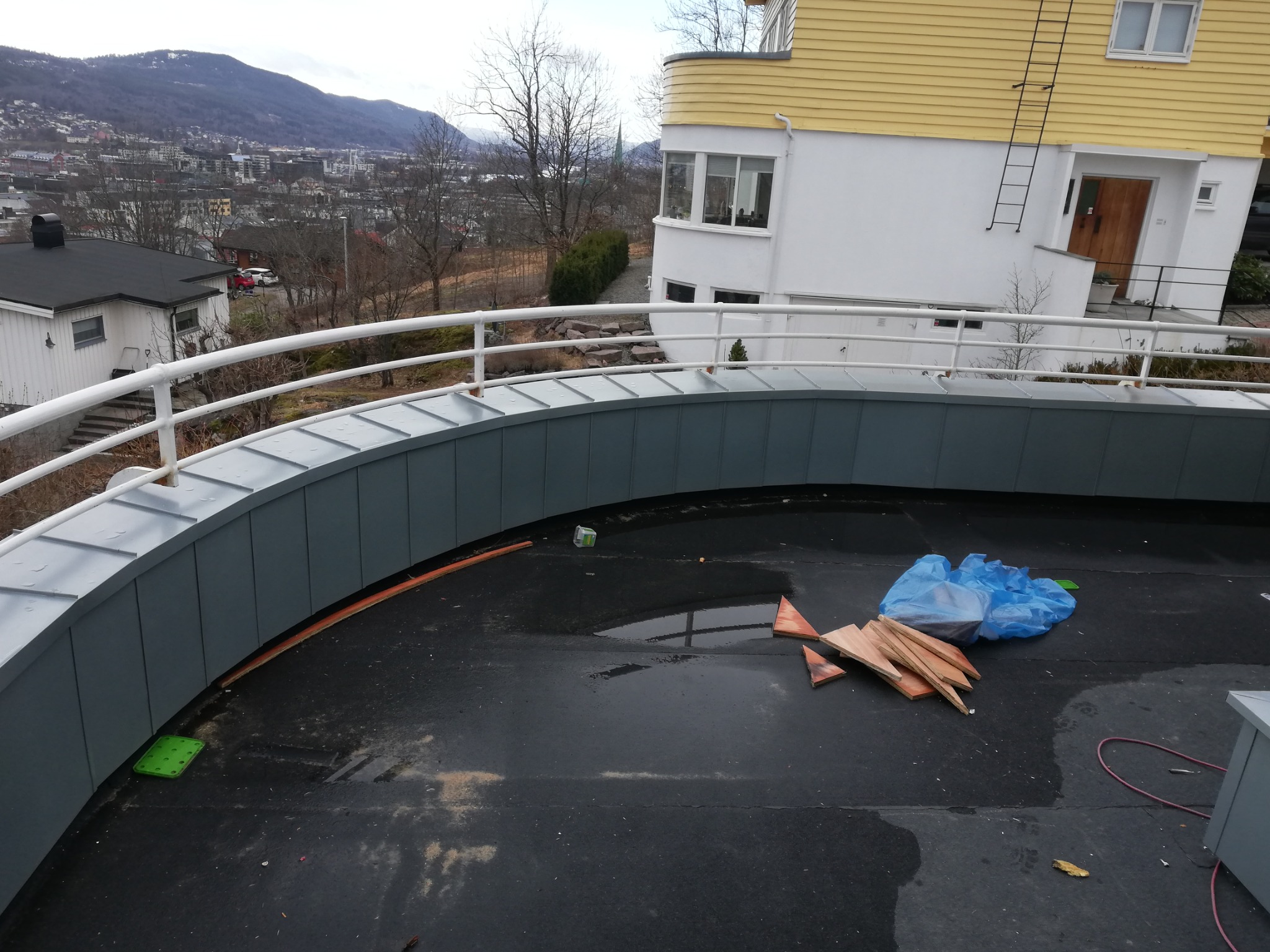 Szara obróbka blacharska na zaokrąglonym dachu z widokiem na miasto, fragmenty drewna i niebieska folia na mokrej powierzchni dachu.