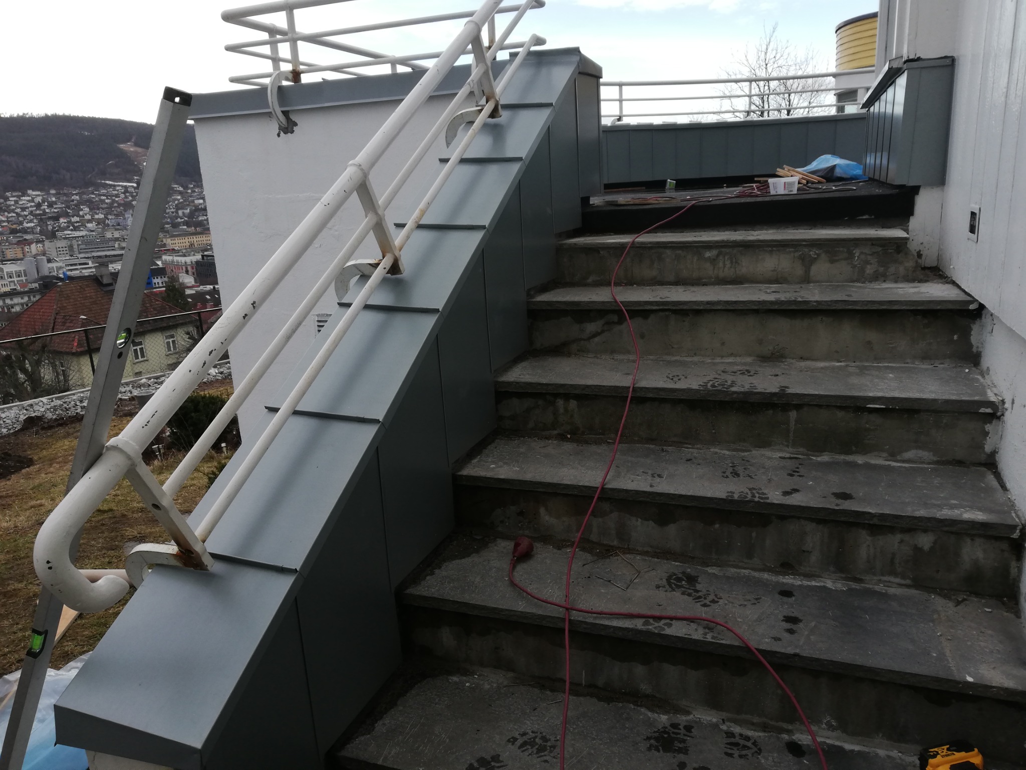 Szare, metalowe wykończenie schodów zewnętrznych z białą balustradą, widok na miasto w tle, przewód elektryczny na schodach.