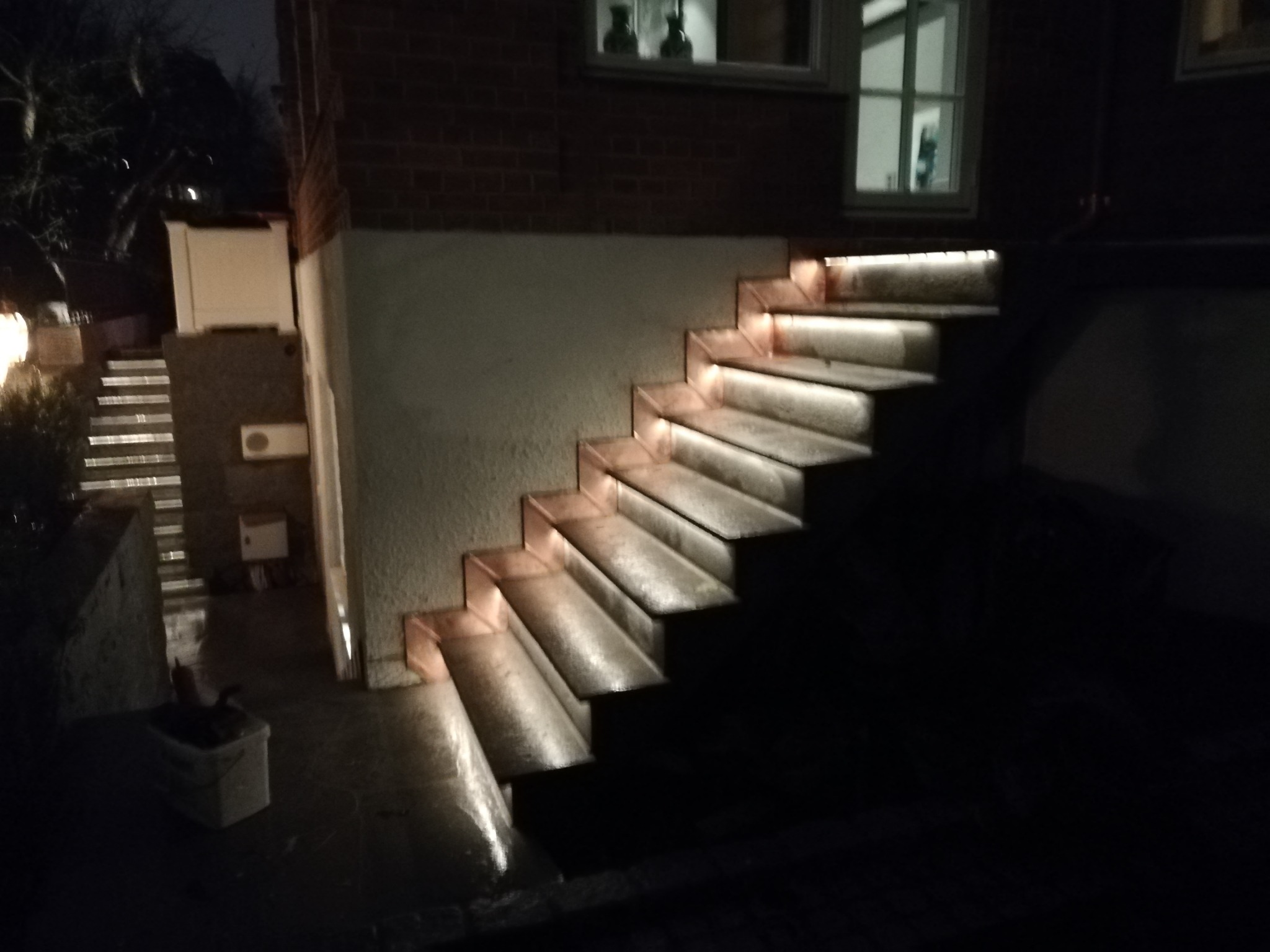 Schody zewnętrzne z podświetleniem LED zamontowanym pod każdym stopniem, prowadzące do budynku z cegły, widoczne okna w górnej części kadru, oświetlenie nocne.