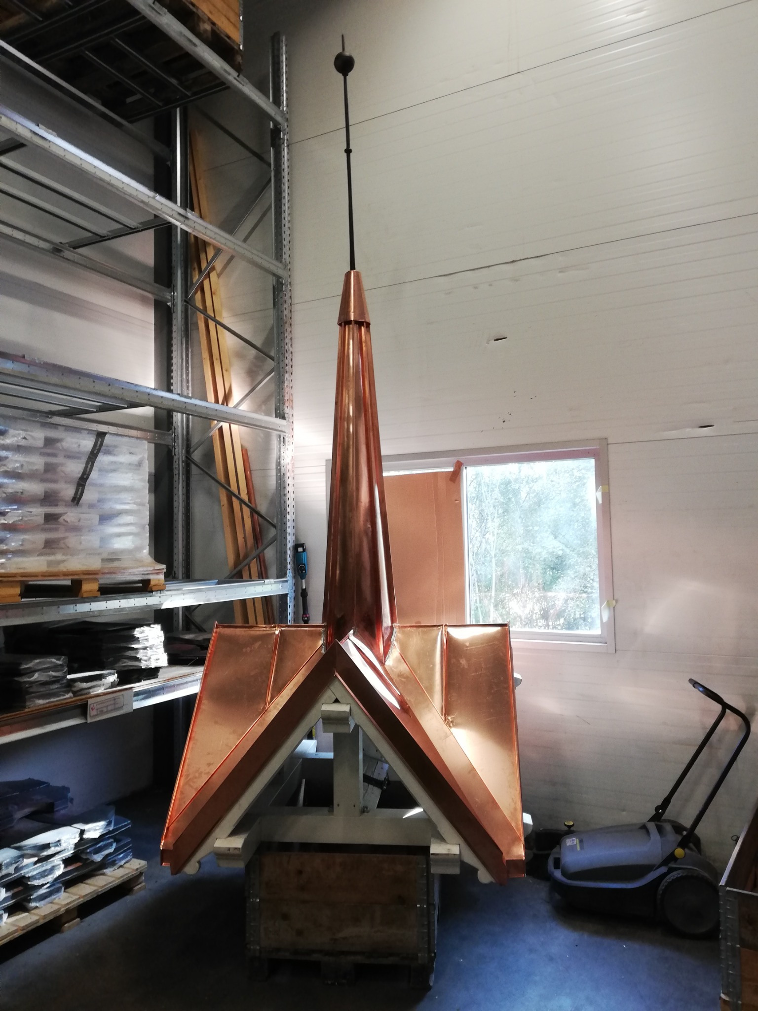 Miedziana iglica dachowa z odgromnikiem, przygotowana do montażu, widoczna w warsztacie na tle regałów z materiałami dekarskimi i okna.