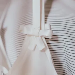 Delikatny, biały top z satynową kokardką na ramiączku, zawieszony na wieszaku z tkaniny w czarno-białe paski.