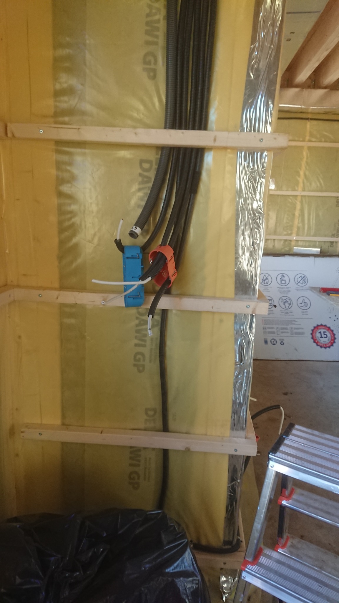 Wewnętrzna instalacja elektryczna w budynku w trakcie budowy: kable w peszlach przymocowane do ściany z izolacją, widoczne puszki instalacyjne, drabina aluminiowa w tle.