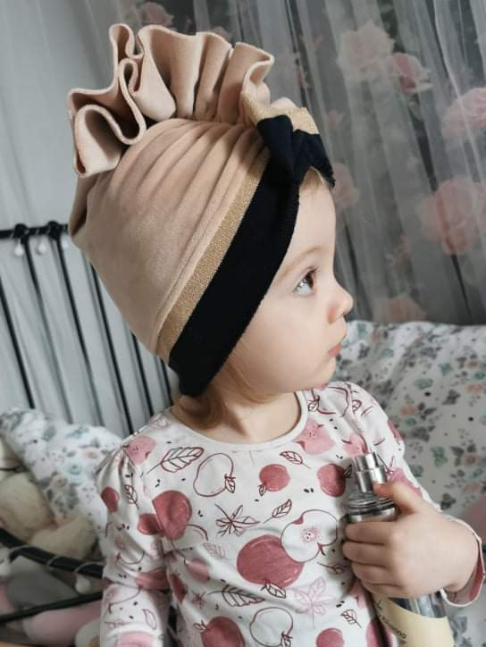 Dziewczynka w beżowym turbanie z falbanami i czarną opaską z złotymi paskami, trzymająca srebrny atomizer na tle pokoju dziecięcego.