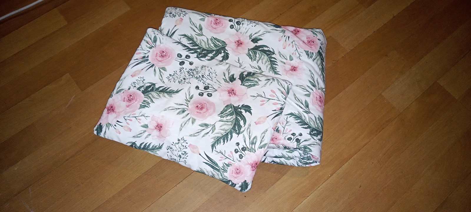 Złożony komplet pościeli dziecięcej z motywem róż i liści na drewnianej podłodze.