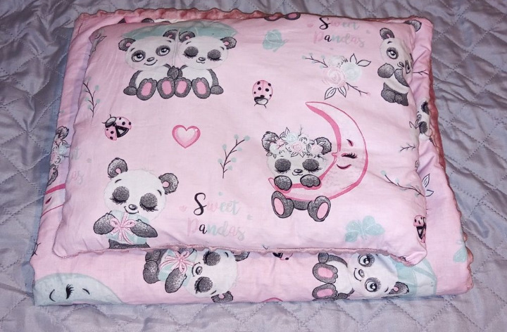 Różowy komplet pościeli dziecięcej z motywem pand, leżący na szarym, pikowanym kocu.
