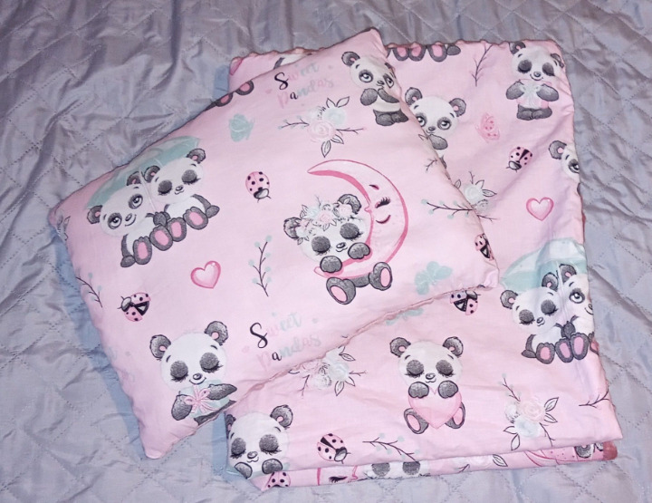 Różowa pościel dziecięca z motywem pand, księżyca i serduszek, ułożona na szarym pikowanym materiale.