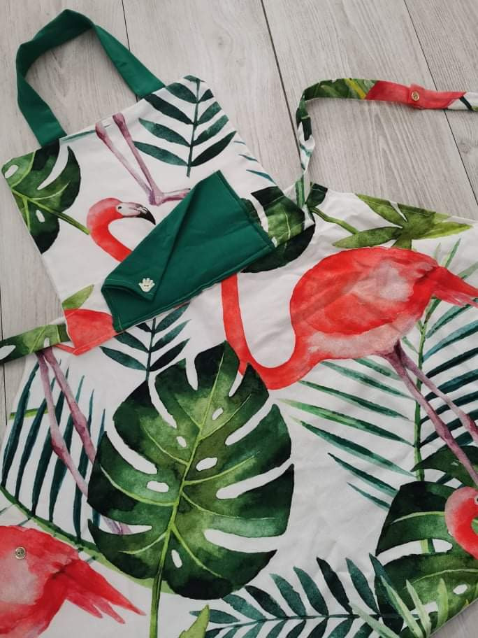 Torba shopper z motywem flamingów i liści tropikalnych, wykończona zielonymi uchwytami i podszewką, leży na drewnianej podłodze.