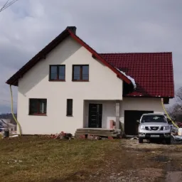 Dwukondygnacyjny dom z czerwonym dachem i białymi ścianami, z samochodem zaparkowanym obok garażu i stertą cegieł przed wejściem, w pochmurny dzień.