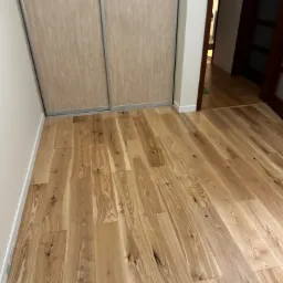 Ułożone panele podłogowe z jasnego drewna z widocznymi słojami, przylegające do ściany i zabudowy przesuwnej szafy, widok na otwarte drzwi do innego pomieszczenia w tle.