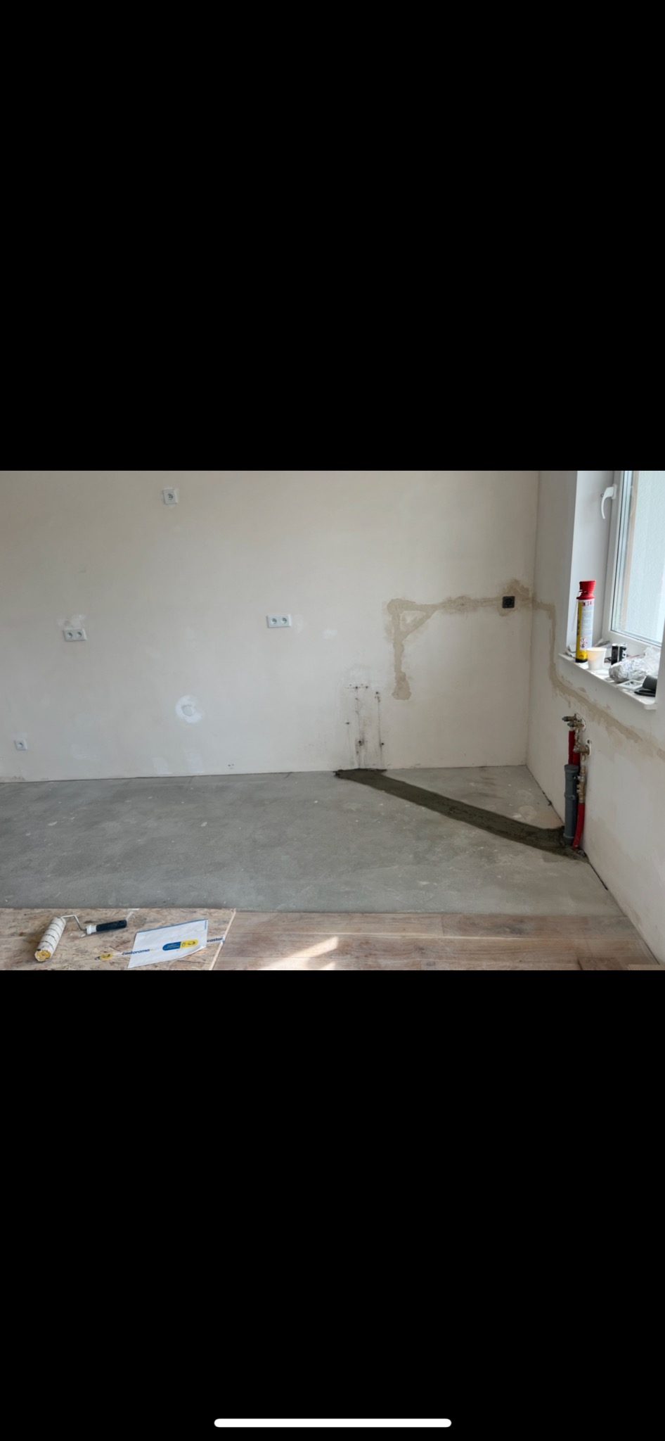 Przygotowanie do prac wykończeniowych: surowa ściana z widocznymi instalacjami elektrycznymi i śladami po naprawach, częściowo wylana podłoga i narzędzia malarskie na panelach podłogowych w rogu...