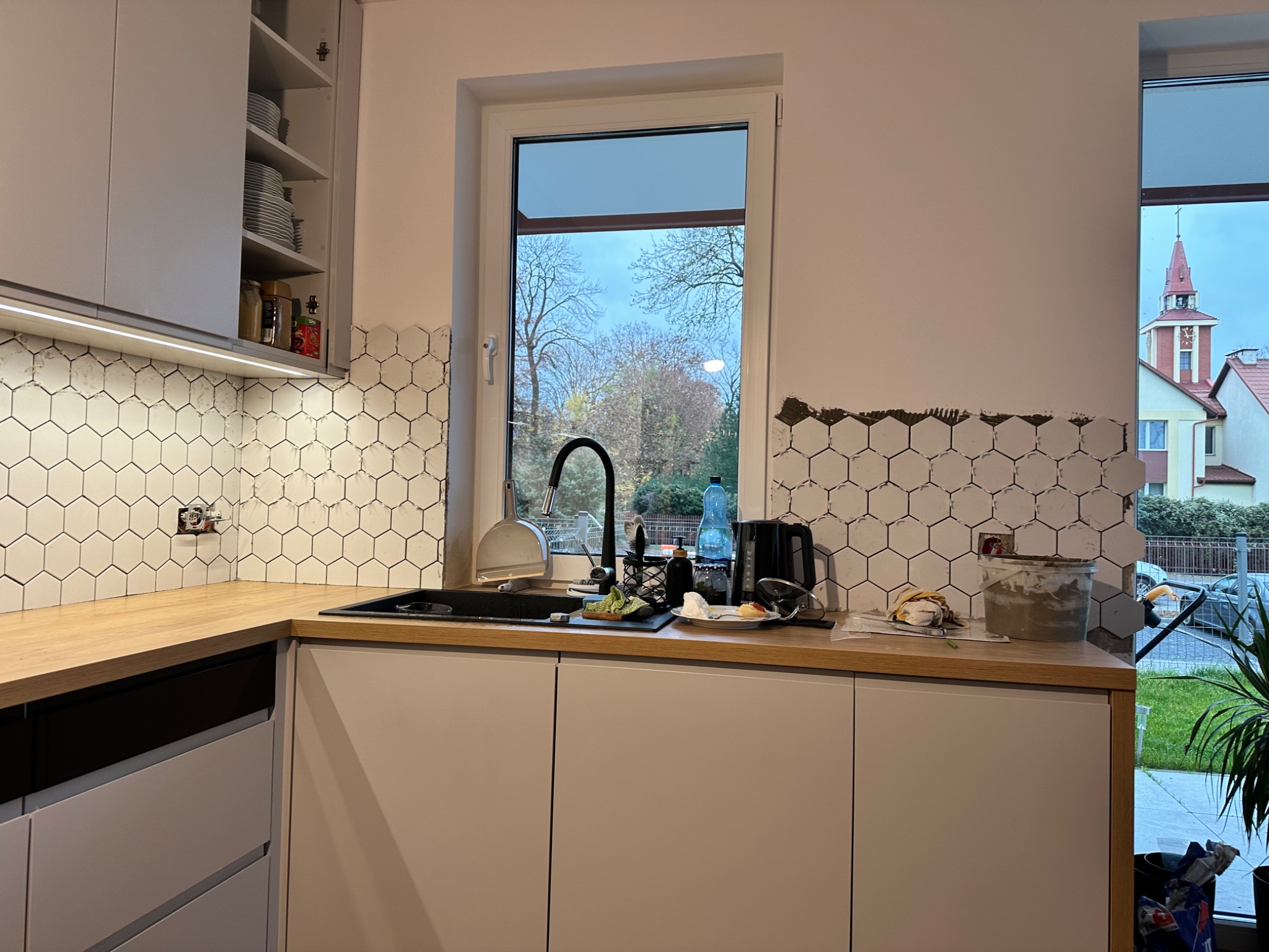 Montaż heksagonalnych płytek ściennych w kuchni, widoczny fragment niedokończonej ściany, fuga w wiadrze i narzędzia, okno z widokiem na kościół w tle.