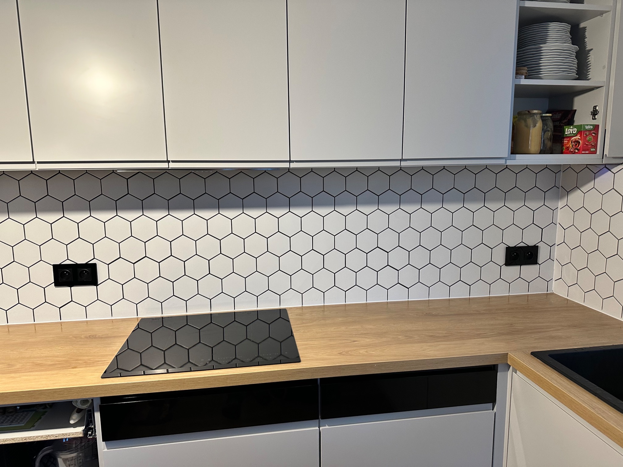 Nowoczesna kuchnia z białymi szafkami, drewnianym blatem i heksagonalnymi płytkami na ścianie, w której zamontowano czarną płytę indukcyjną.