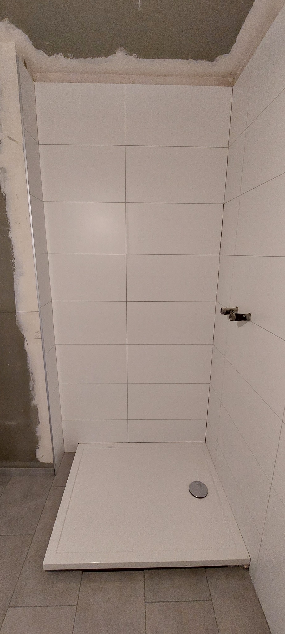 Narożnik kabiny prysznicowej z zamontowanym brodzikiem i częściowo wyłożonymi białymi płytkami, widoczne niedokończone prace przy suficie i ścianie obok.